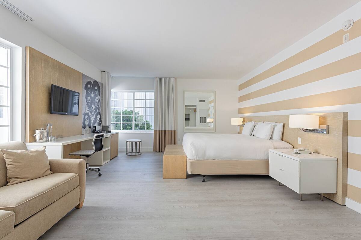 Ein Hotelzimmer mit Bett, Schreibtisch, Stuhl, Sofa, Fernseher und einem Fenster mit Vorhängen.