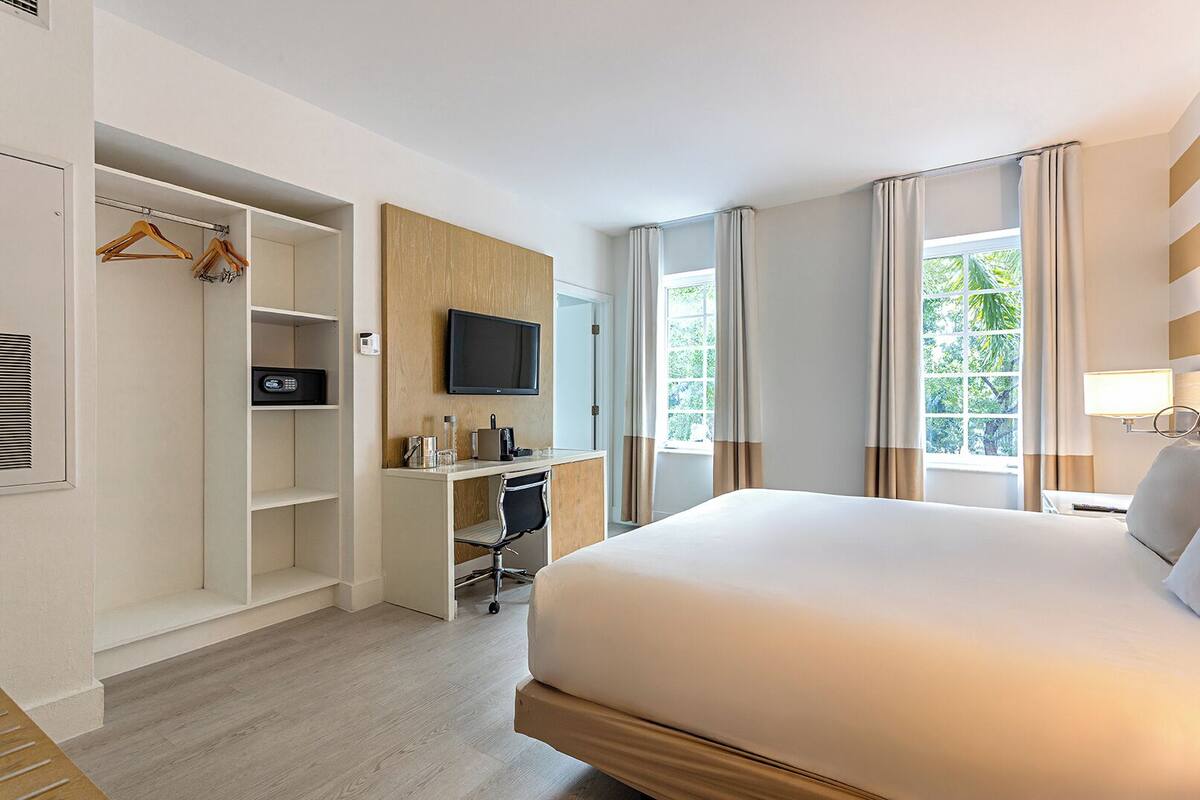 Ein Hotelzimmer mit Bett, Schreibtisch mit Stuhl, Fernseher, Kleiderschrank und einem Fenster mit Vorhängen.