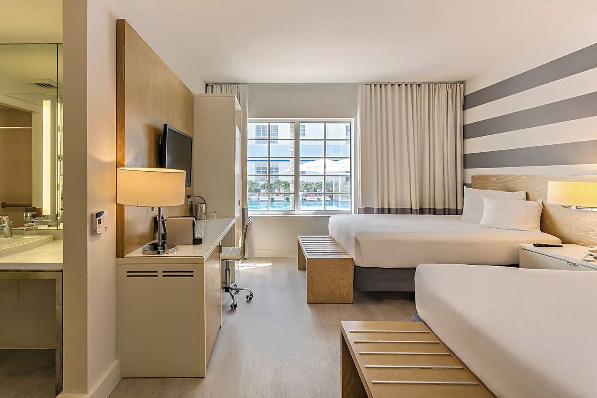 Ein Hotelzimmer mit Bett, Nachttisch, Lampe, Schreibtisch mit Stuhl und einem Fenster mit Vorhängen.