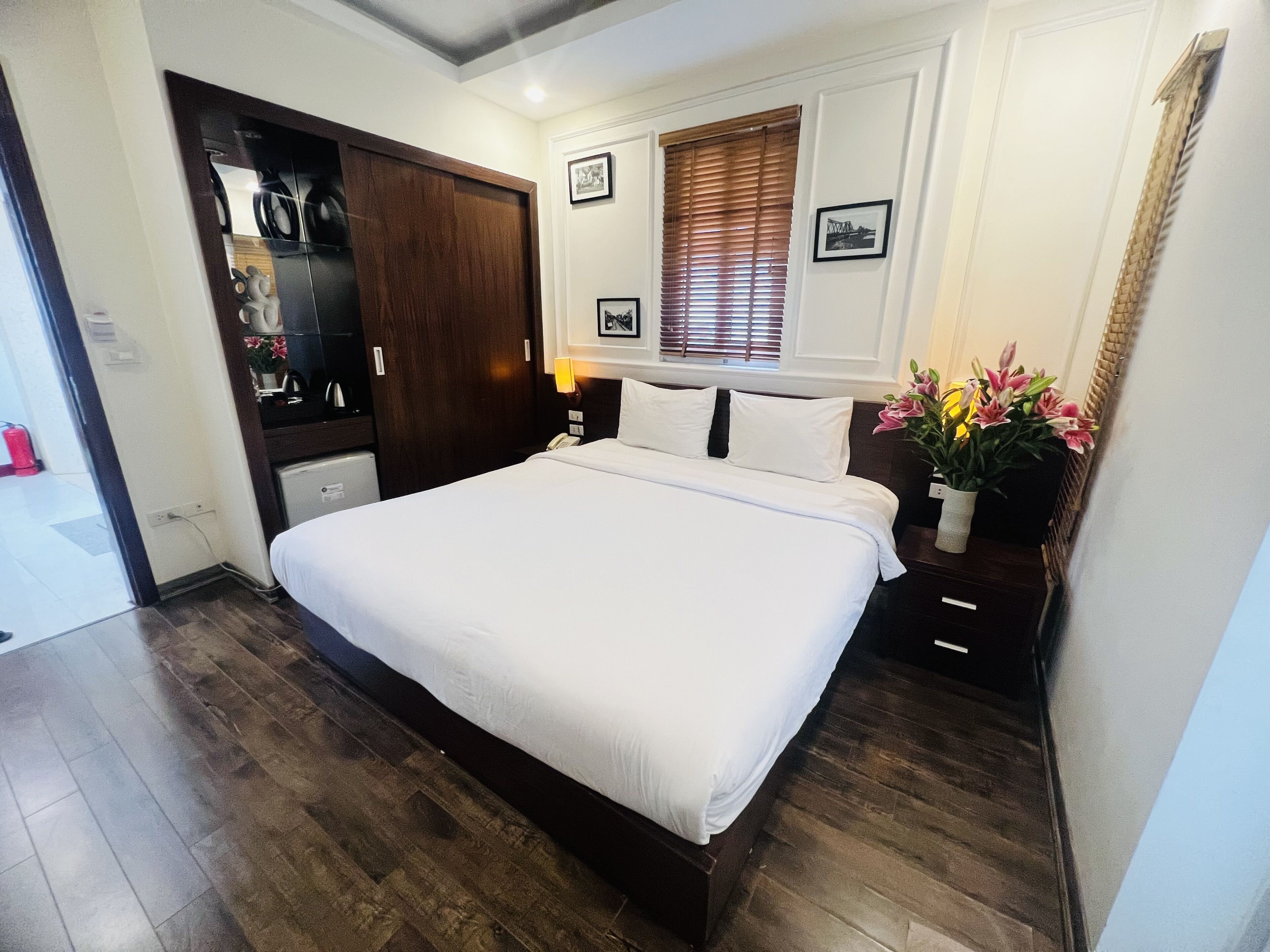 A neatly arranged bedroom with a large bed, wooden wardrobe, and a small bedside table with a lamp.