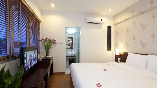 A hotel room with a bed, a TV on a wooden desk, a bathroom with a sink and mirror, and a wall-mounted air conditioner. - Rising Dragon Legend Hotel (Hanoi)