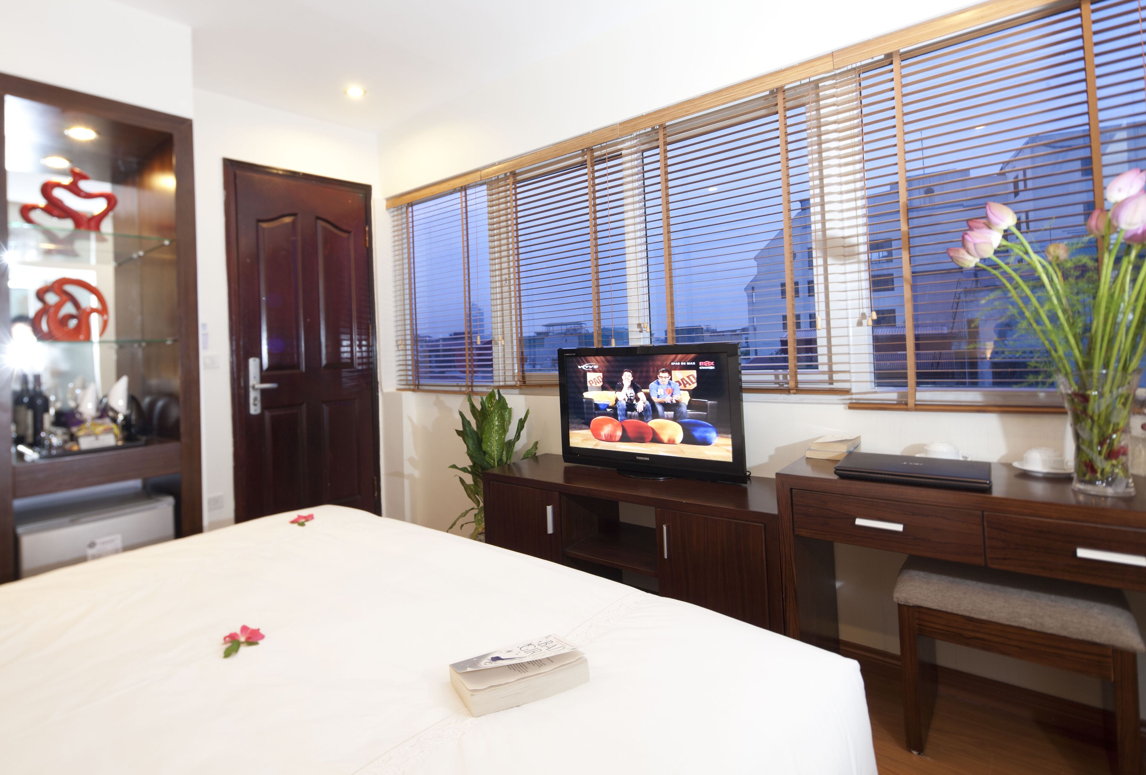 A hotel room with a bed, a desk with a laptop, a television displaying a sports event, and a window with blinds.