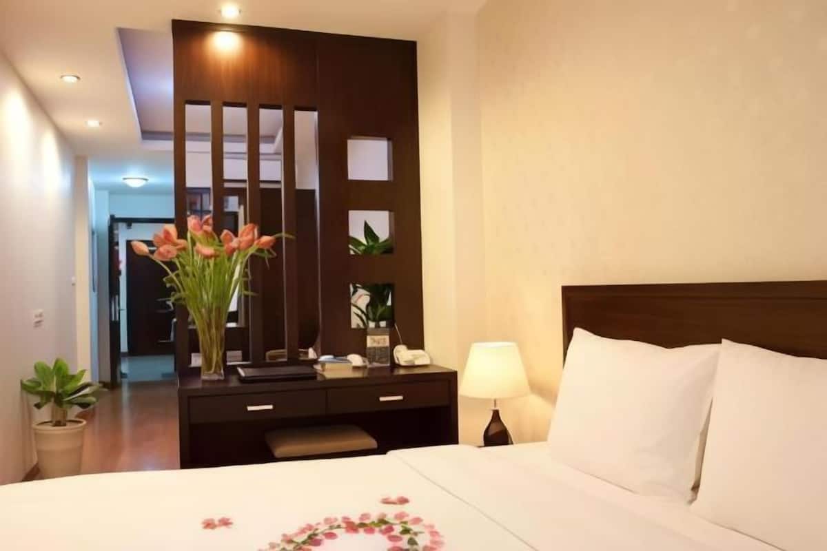 A hotel room with a bed, bedside table, lamp, and a desk with a mirror and plants.