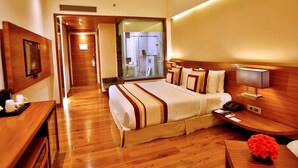 A hotel room with a wooden floor, a bed with a white and brown striped bedspread, a bedside table with a lamp, a small table with a vase of flowers, and a mirror on the wall. - Park Ascent (Noida)