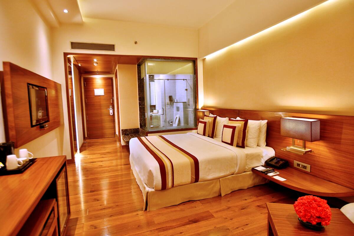 A hotel room with a wooden floor, a bed with a white and brown striped bedspread, a bedside table with a lamp, a small table with a vase of flowers, and a mirror on the wall.