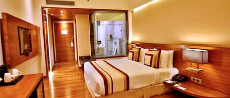 Habitación de hotel con piso de madera, una cama con colcha a rayas blancas y marrones, una mesita de noche con lámpara, una pequeña mesa con un jarrón de flores y un espejo en la pared.