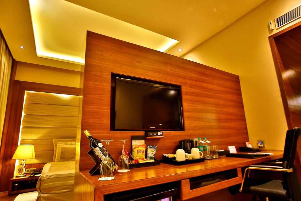 A hotel room with a large flat-screen TV mounted on a wooden wall, a bedside table with a lamp, a minibar, and a seating area with a chair and a small table.