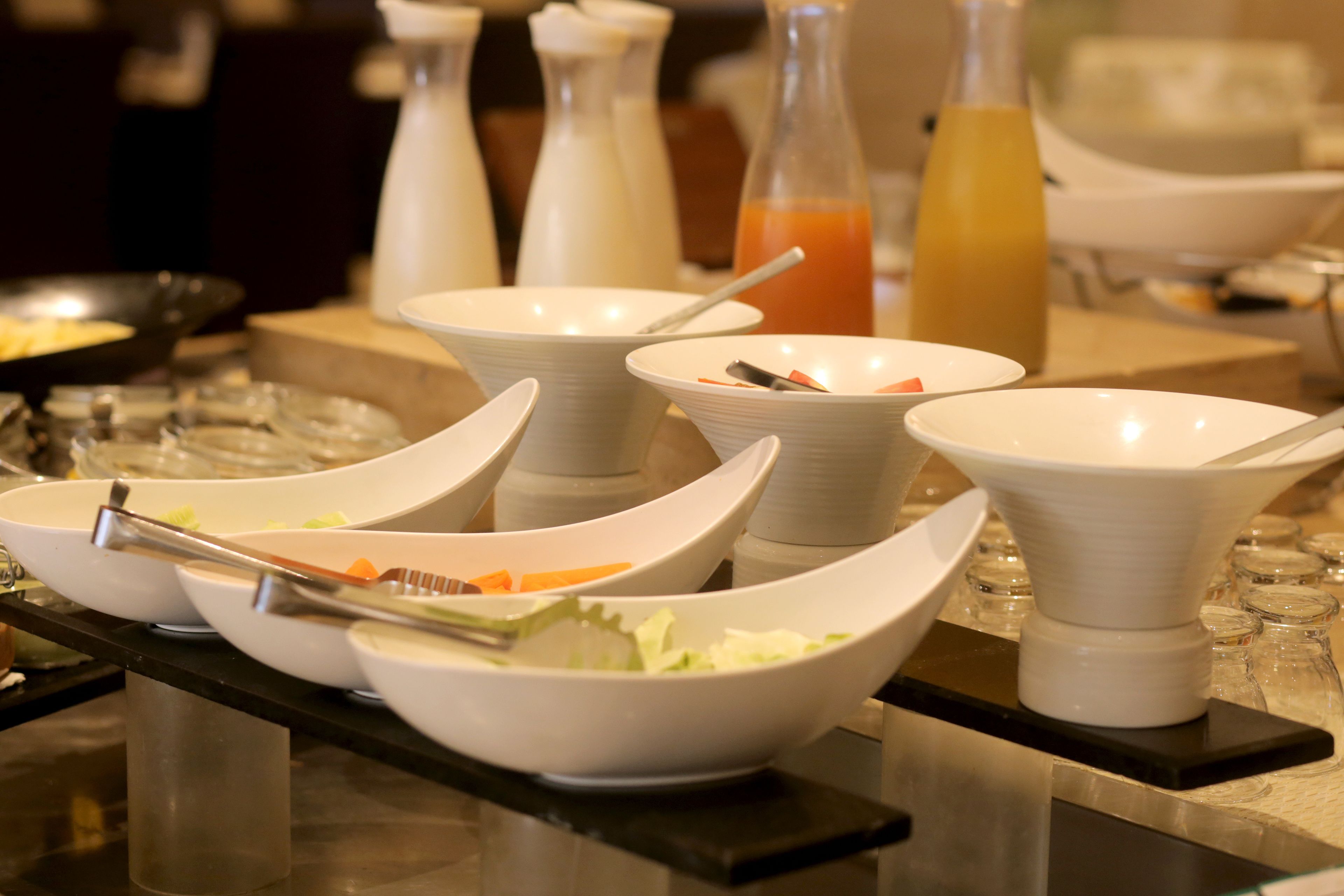 Una mesa de buffet con varias fuentes blancas llenas de comida, una botella de jugo de naranja y una jarrón de agua.