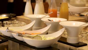 Una mesa de buffet con varias fuentes blancas llenas de comida, una botella de jugo de naranja y una jarrón de agua.