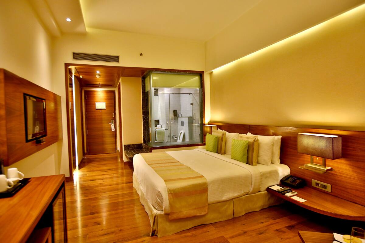 A hotel room with a wooden floor, a bed with a white and beige bedspread, a bedside table with a lamp, and a bathroom visible through an open door.
