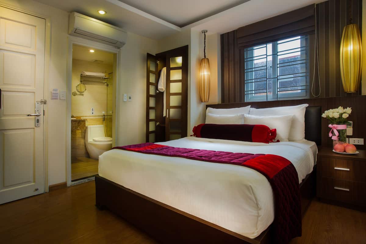 A hotel room with a large bed, a wooden floor, a bathroom with a toilet and sink, and a window with curtains.