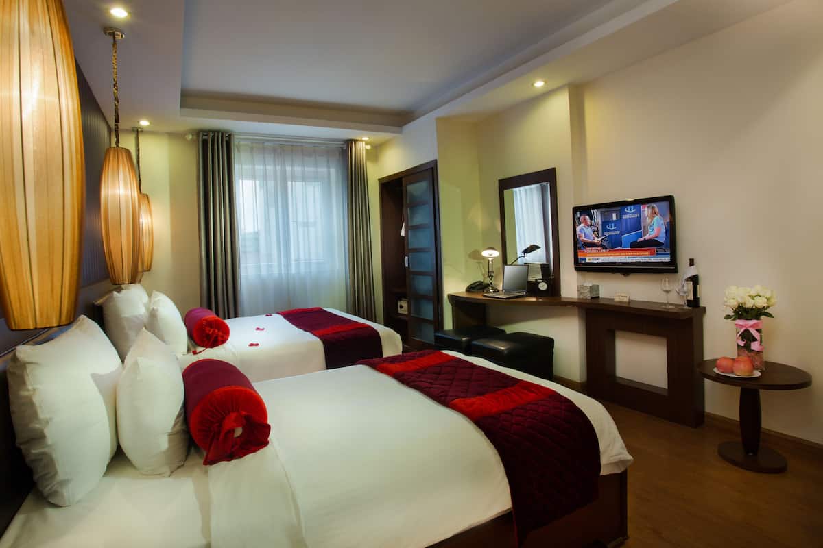 A hotel room with two beds, a desk with a computer, a television, and a small table with a vase of flowers.