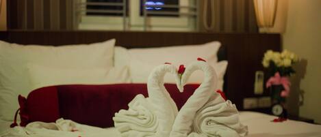 Habitación de hotel con cama que incluye un cabecero rojo, ropa de cama blanca y dos toallas con forma de cisne adornadas con pétalos de rosa.