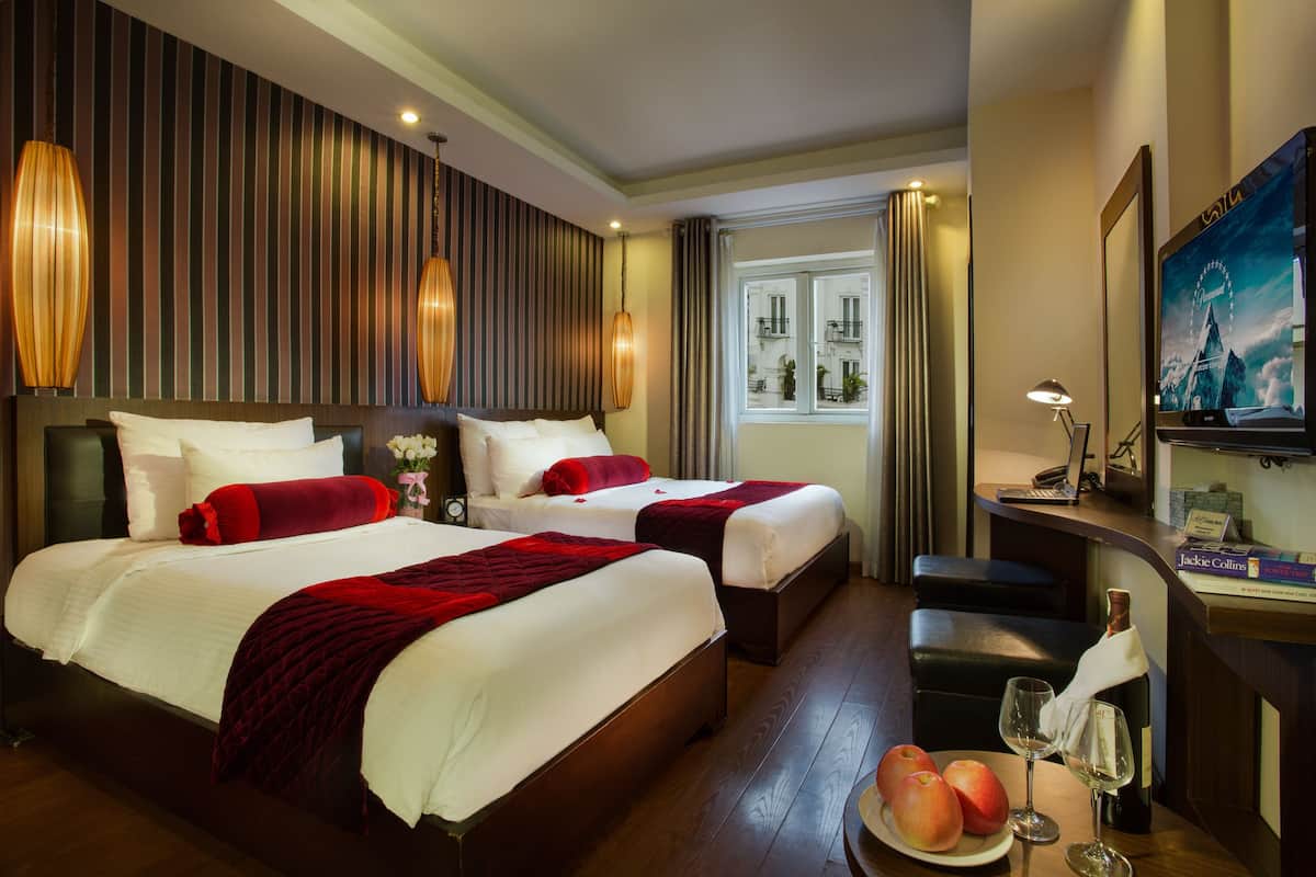 A hotel room with two beds, a desk with a computer, a TV, and a window with a view of buildings.