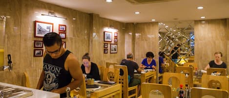 A restaurant interior with wooden tables and chairs, framed pictures on the walls, and a person serving at the counter.