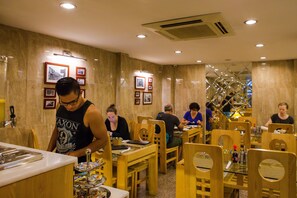 A restaurant interior with wooden tables and chairs, framed pictures on the walls, and a person serving at the counter. - Golden Art Hotel (Hanoi)