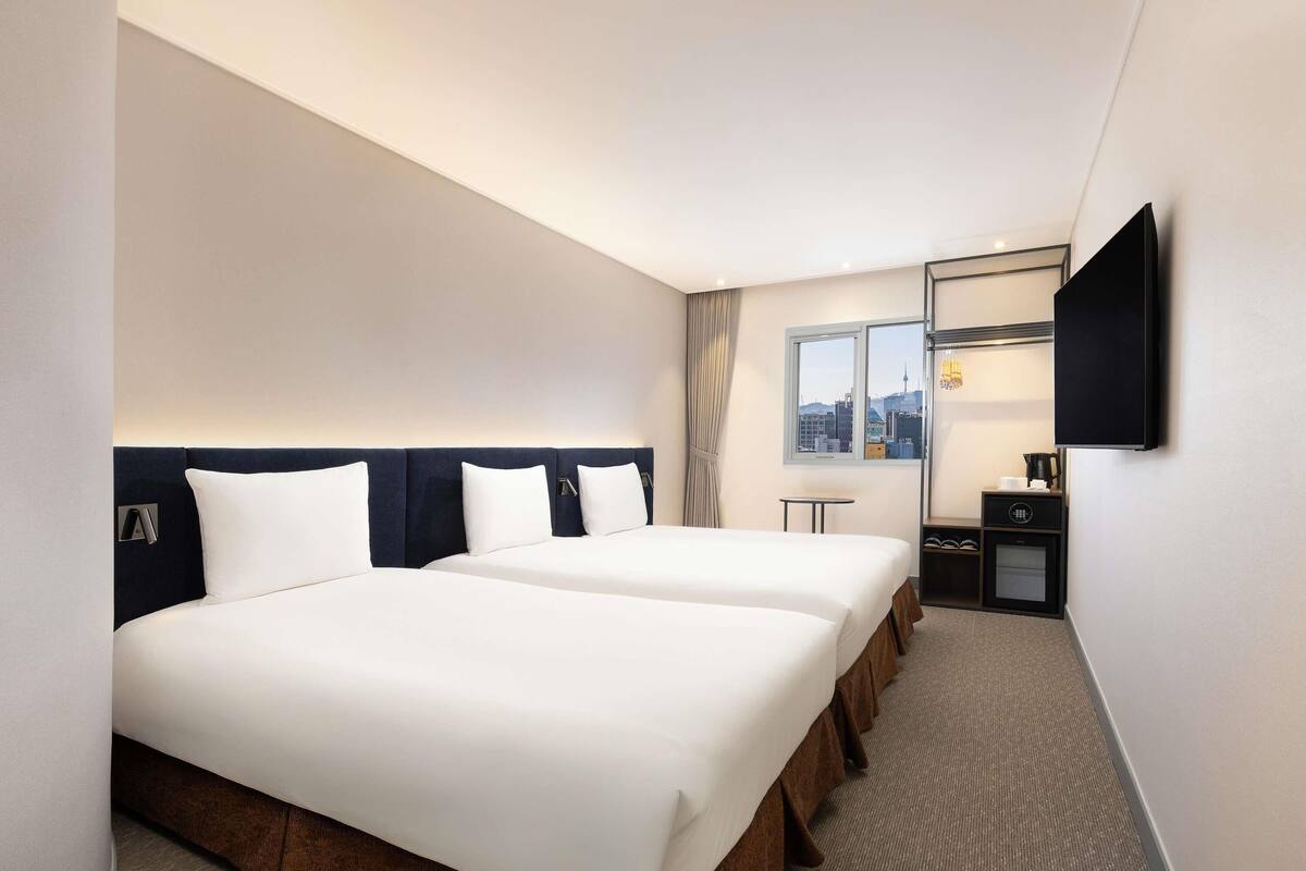 Habitación de hotel con dos camas, televisión, una mesita y ventana con vista a la ciudad.