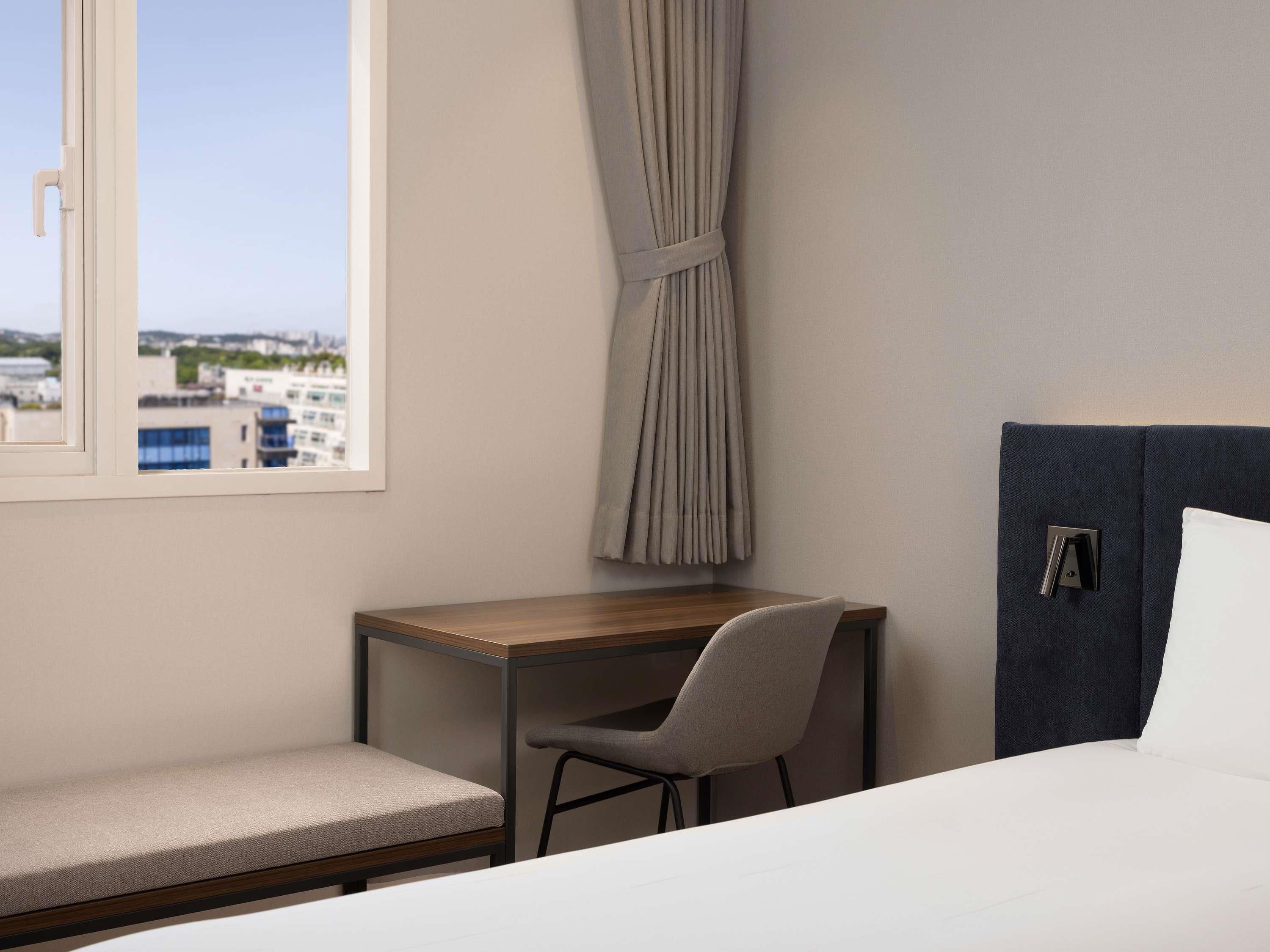 A hotel room with a bed, a desk with a chair, a view of buildings through a window, and a curtain.