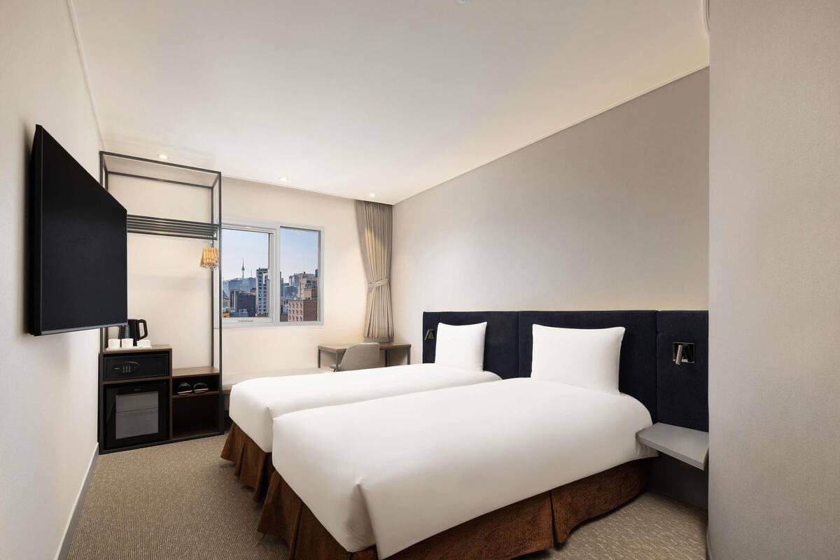Ein Hotelzimmer mit zwei Betten, einem Fernseher, einem Schreibtisch und einem Fenster mit Blick auf die Stadt.