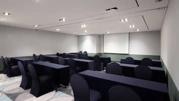 A conference room with multiple tables and chairs, recessed ceiling lights, and a projector screen.