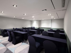 Uma sala de conferências com várias mesas e cadeiras, luminárias embutidas no teto e uma tela de projetor.