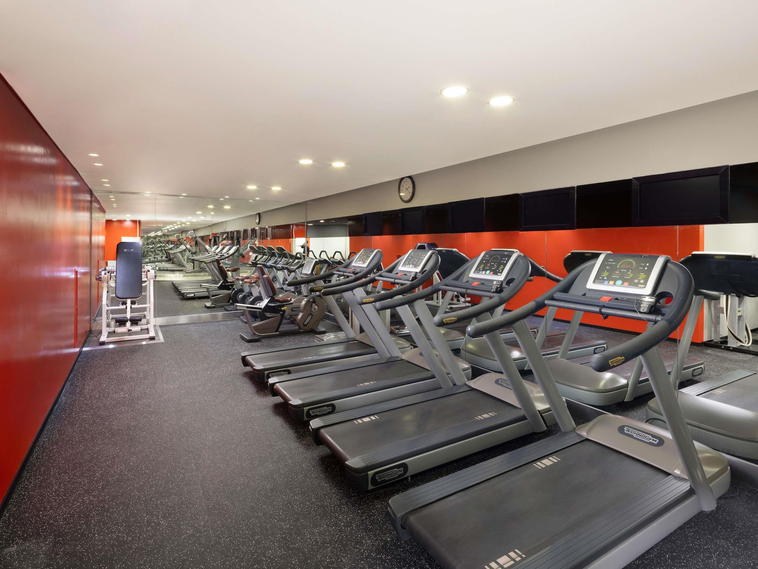 Une salle de sport avec plusieurs tapis de course alignés en rangées, une horloge fixée au mur et un motif mural rouge et noir.