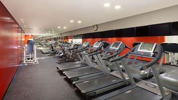 Une salle de sport avec plusieurs tapis de course alignés en rangées, une horloge fixée au mur et un motif mural rouge et noir.
