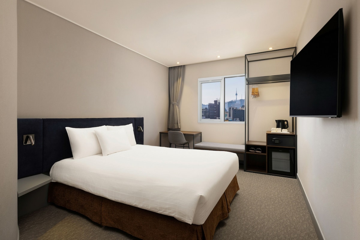 Une chambre d’hôtel équipée d’un lit, d’un bureau, d’une chaise, d’une télévision et d’une fenêtre donnant sur la ville.