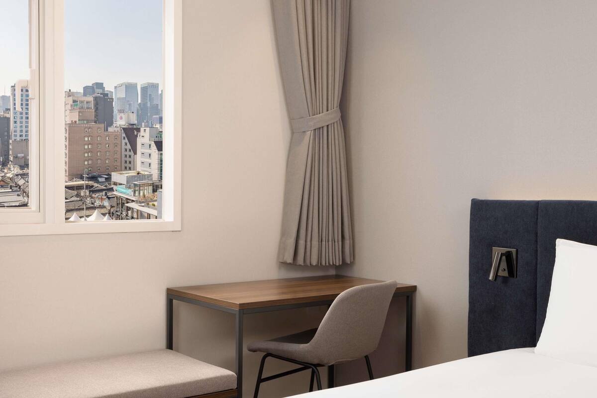 Ein Hotelzimmer mit einem Bett, einem Schreibtisch, einem Stuhl und Blick auf eine Stadtlandschaft, sowie einem Vorhang.