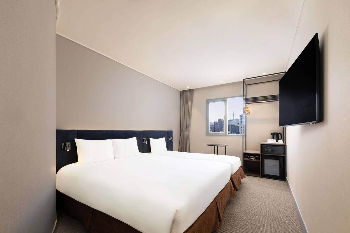 Ein Hotelzimmer mit einem großen Bett, einem an der Wand befestigten Fernseher, einem kleinen Tisch mit Kaffeemaschine und einem Fenster mit Blick auf die Stadt.