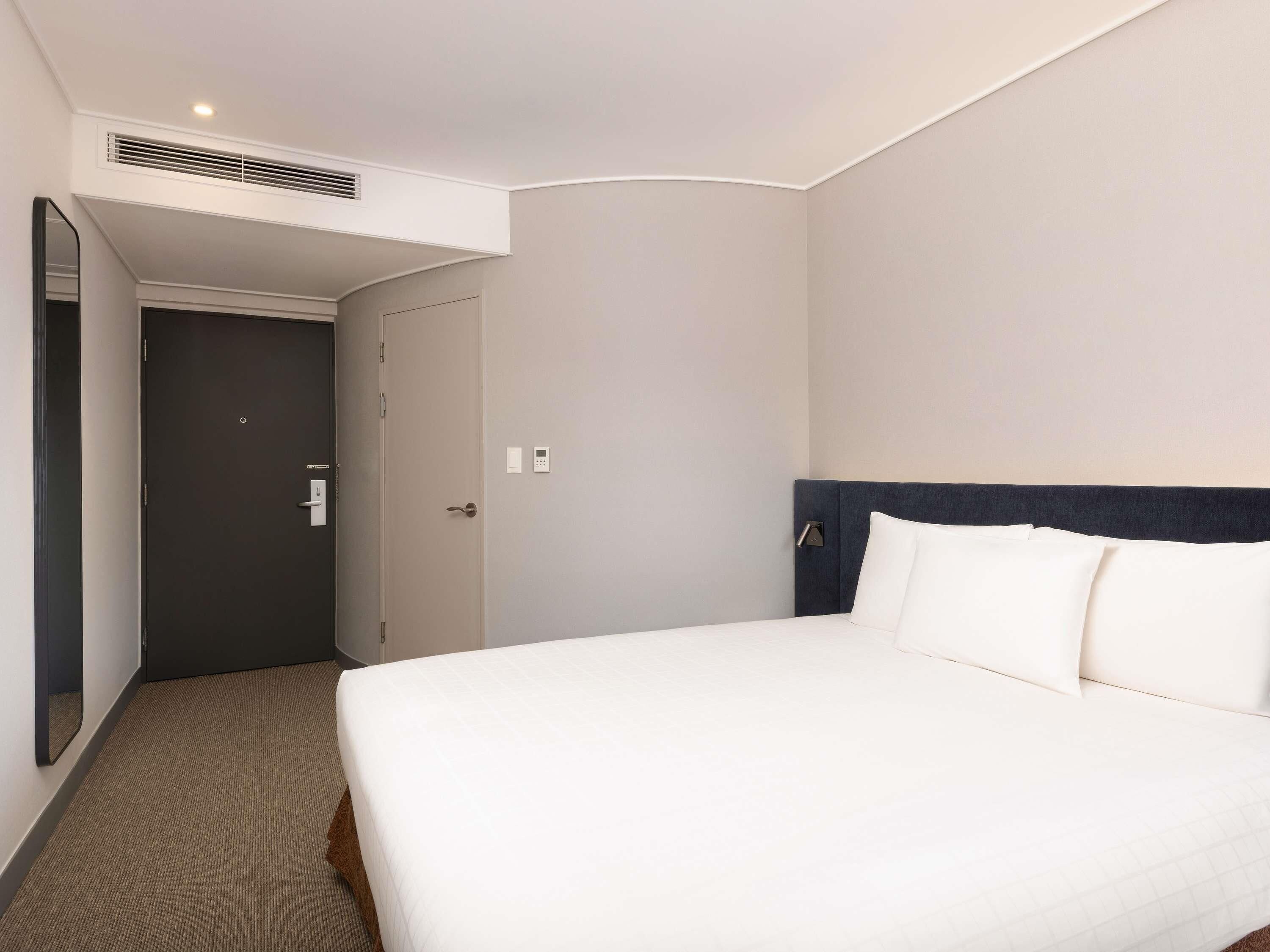Une chambre d’hôtel avec un lit, une porte, un miroir et un panneau de commande fixé au mur.