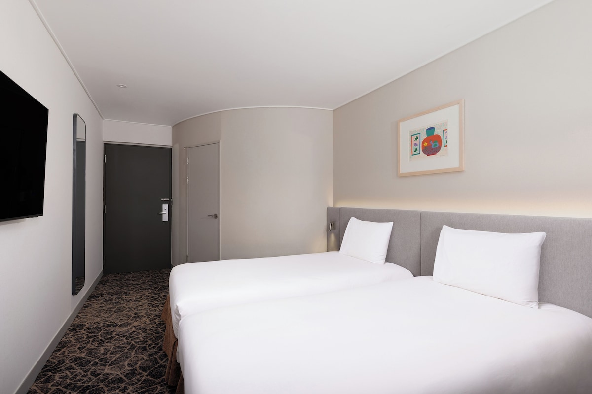 Une chambre d’hôtel avec deux lits, un grand miroir, un tableau encadré au mur et un sol recouvert de moquette.