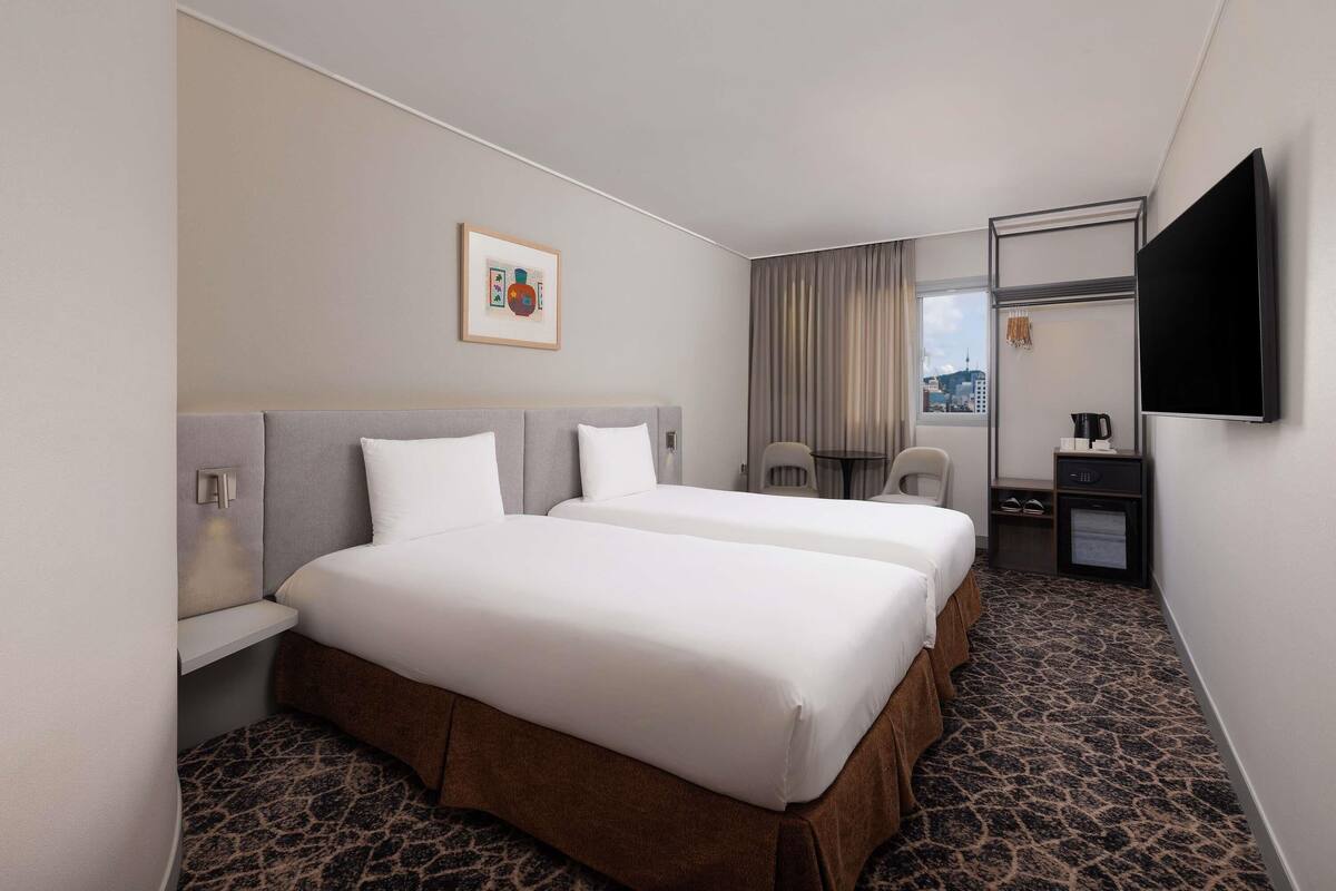Ein Hotelzimmer mit zwei Betten, einem Flachbildfernseher, einem kleinen Tisch mit Stühlen, einer Kaffeemaschine und Blick auf die Stadt.