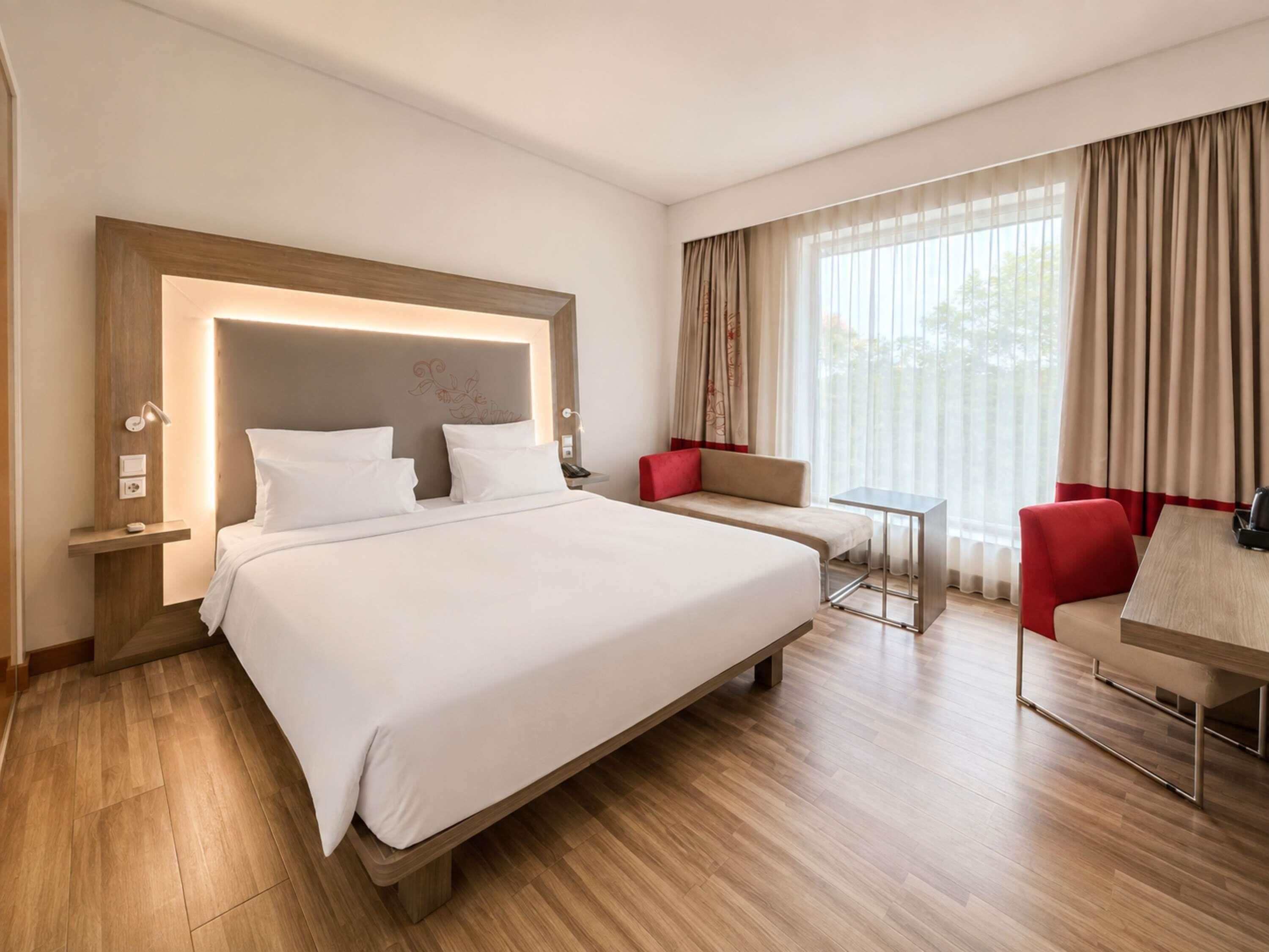 A modern hotel room with a large bed, a seating area with a red chair, and a small table.