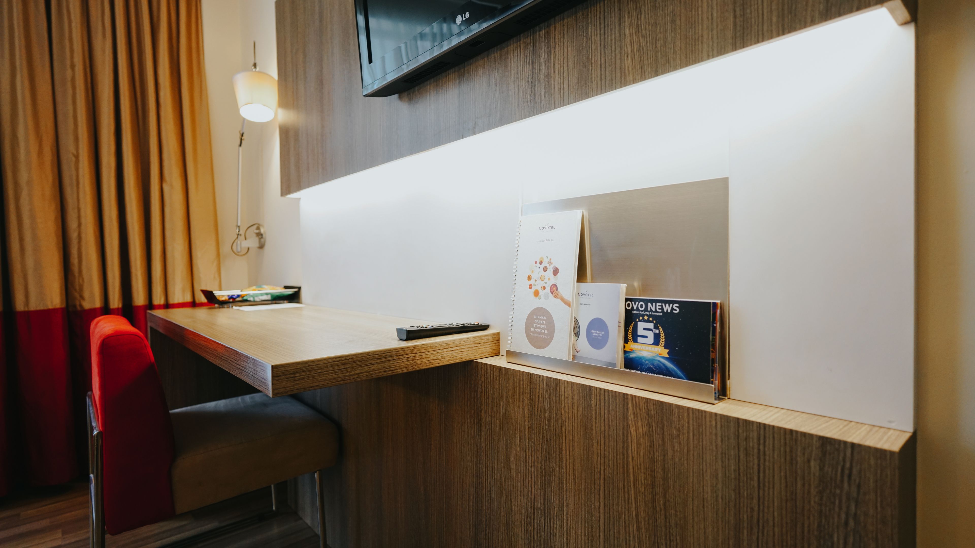 Un comptoir d’accueil d’hôtel avec un comptoir en bois, une chaise rouge, une télécommande et un présentoir à magazines avec un numéro spécial consacré au « 5e anniversaire ».