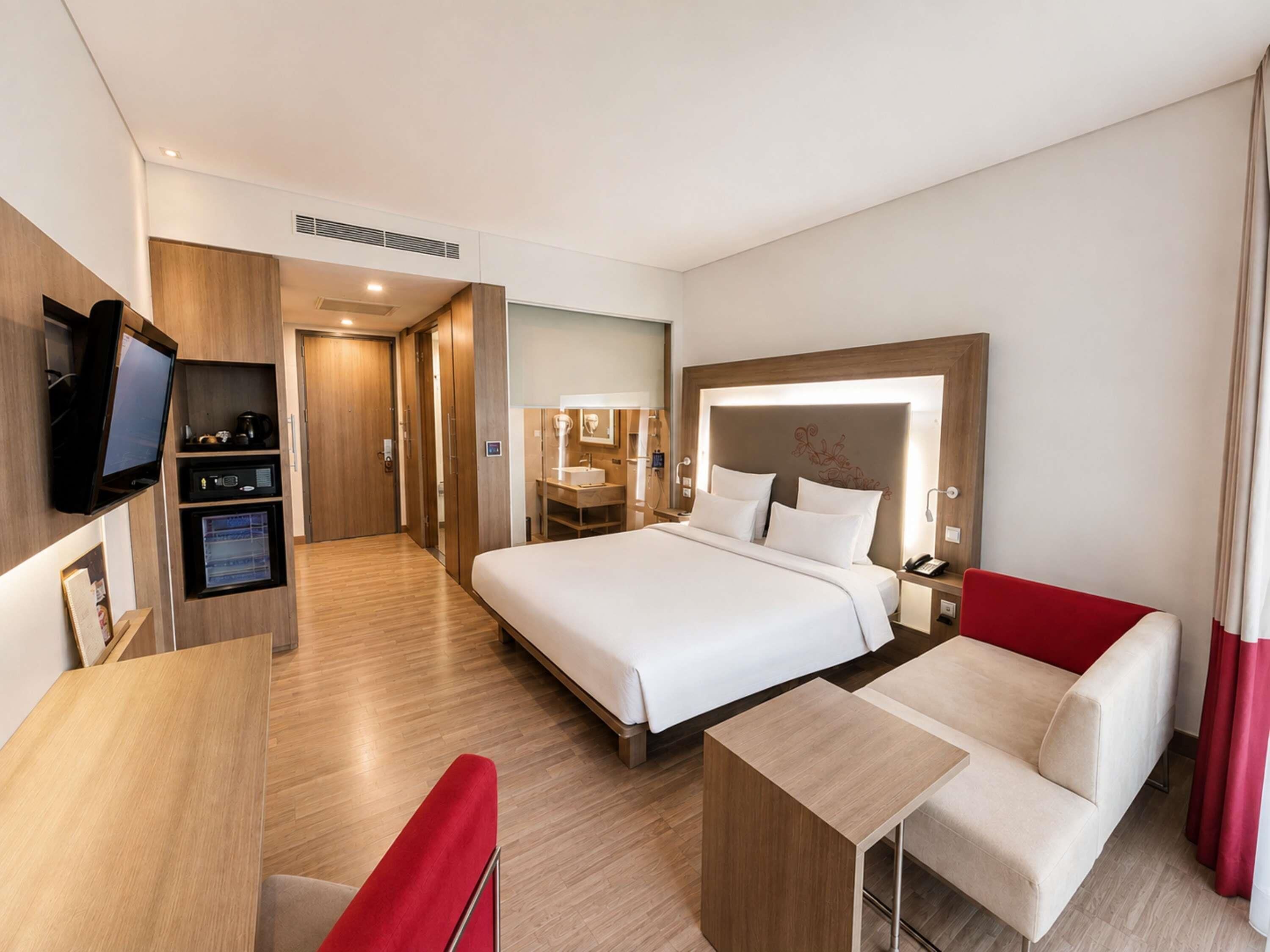 Una habitación de hotel con cama, escritorio, televisor, minibar y una silla roja.