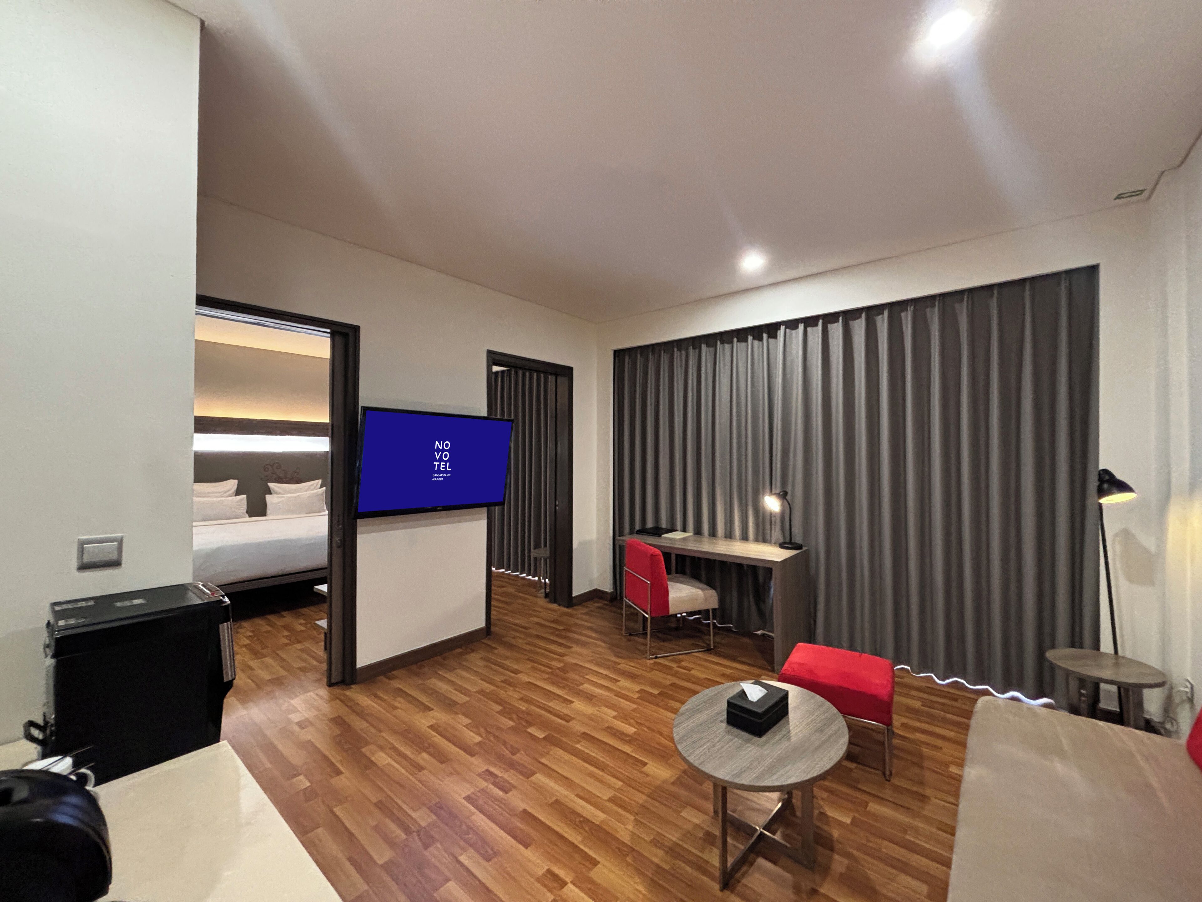 Ein modernes Hotelzimmer mit Holzboden, einem Bett mit weißer Bettwäsche, einem Schreibtisch mit Stuhl, einem kleinen runden Tisch mit einer Schachtel und einem Fernseher an der Wand.