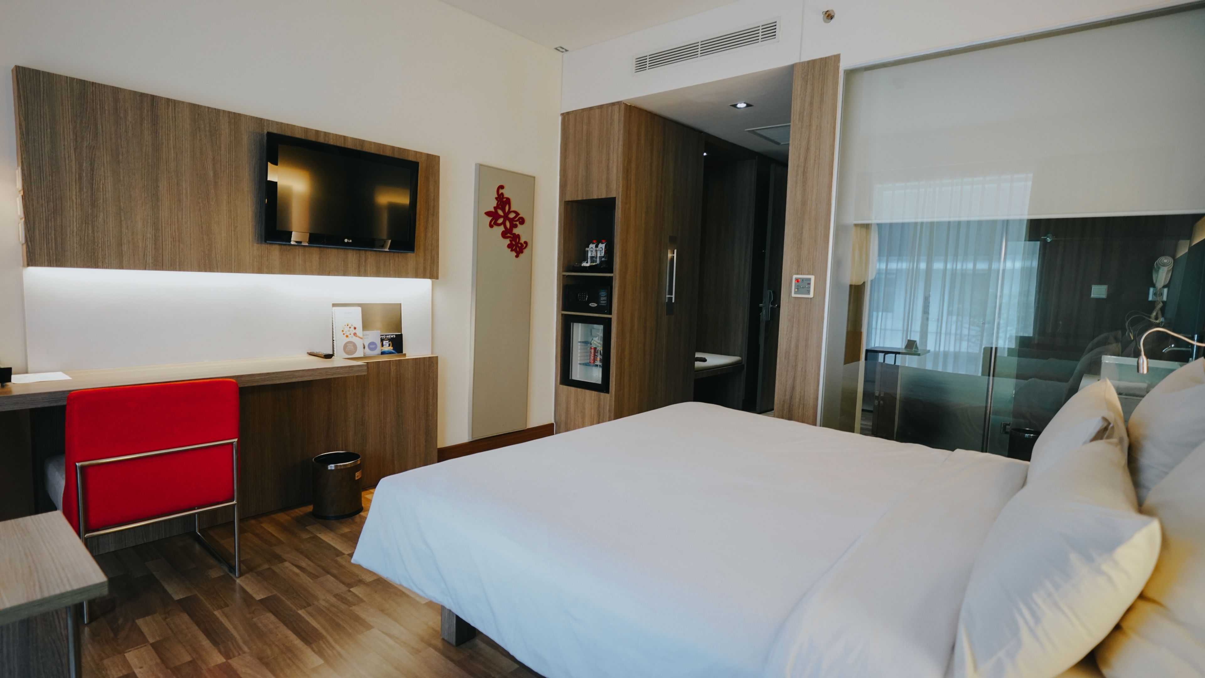 Habitación de hotel con cama, escritorio y una silla roja, televisor montado en la pared y minibar.