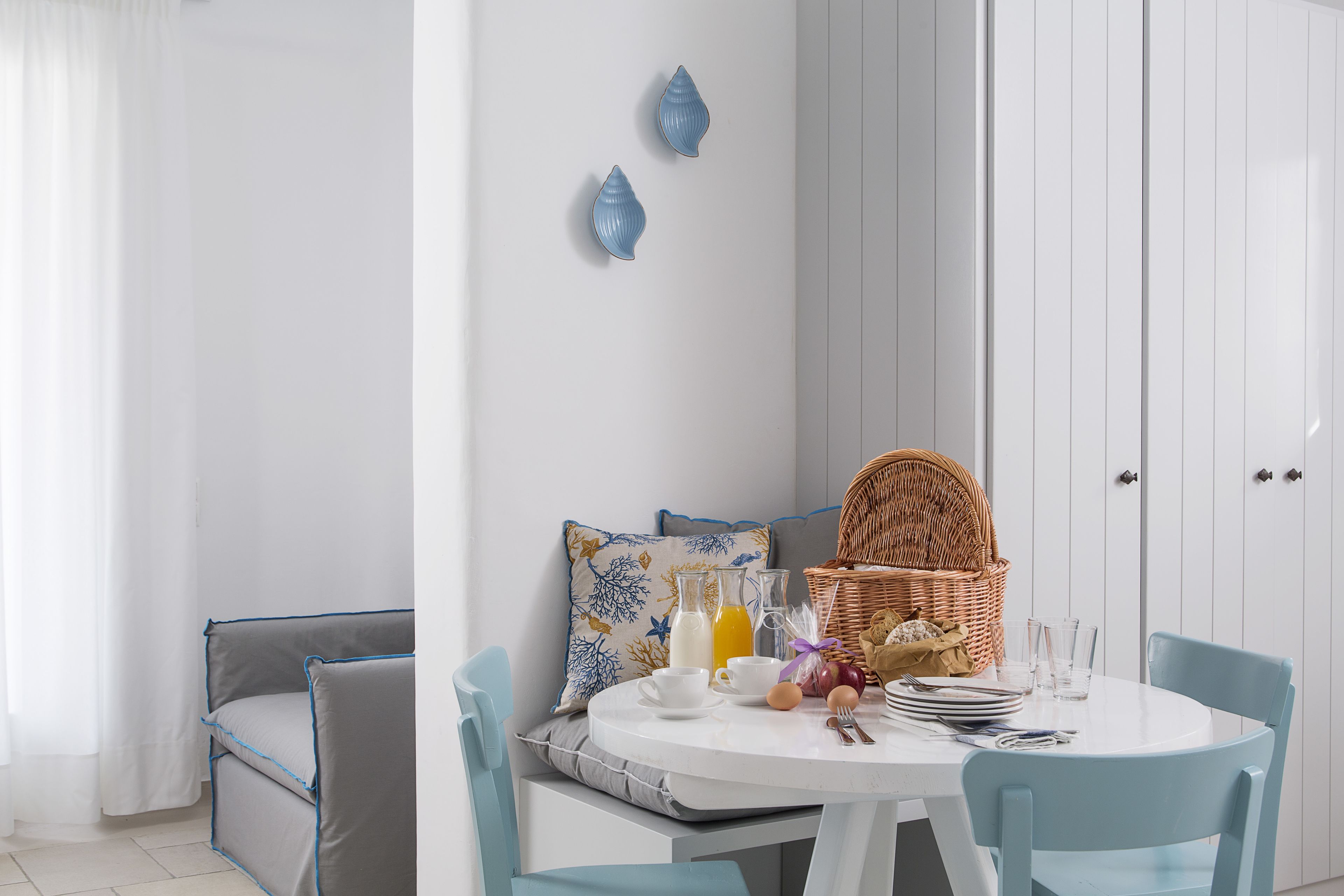 A dining area with a white table, blue chairs, a wicker basket, breakfast items, and a cushioned bench.