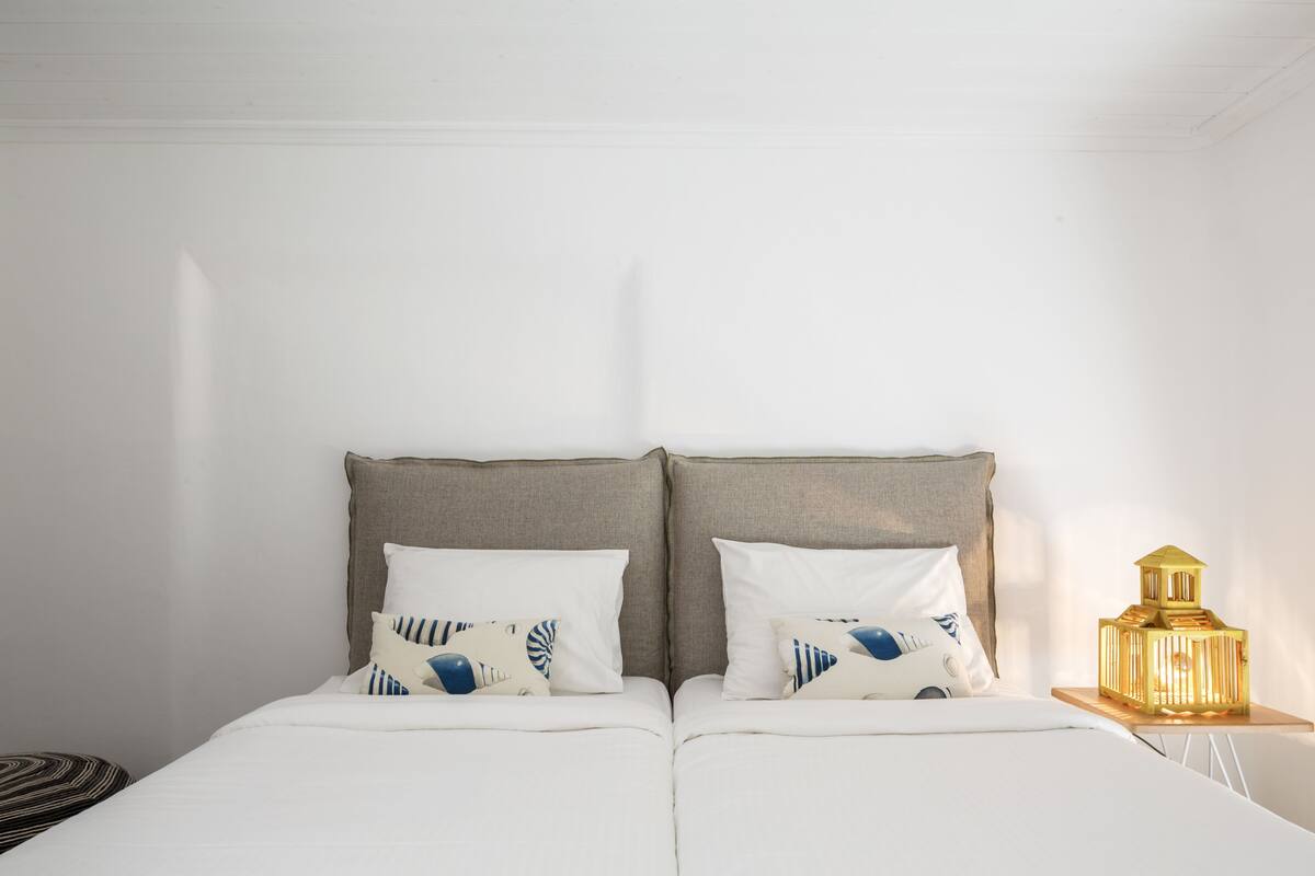 Un lit double avec une tête de lit, deux oreillers, une table de chevet avec une lampe et un coussin rayé posé sur le sol.
