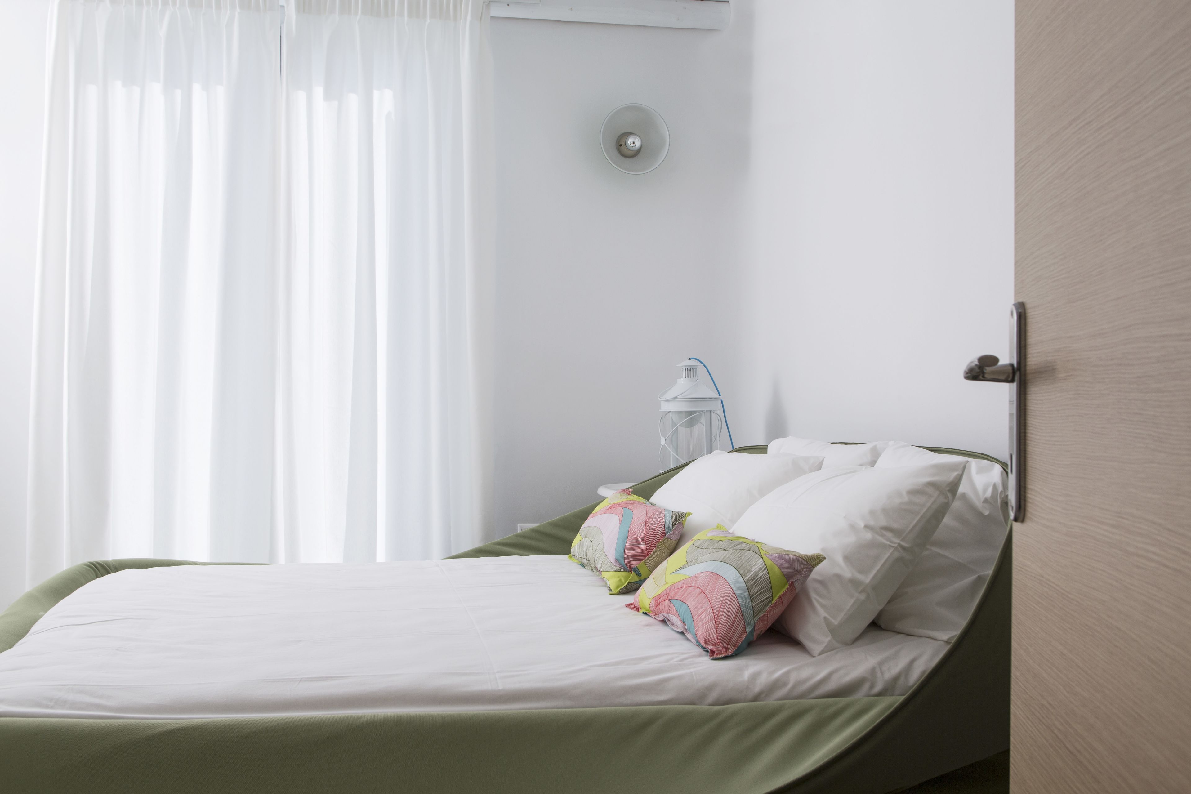 A neatly made bed with a green bedspread and colorful pillows, a white bedside table with a lamp, and a window with sheer curtains.