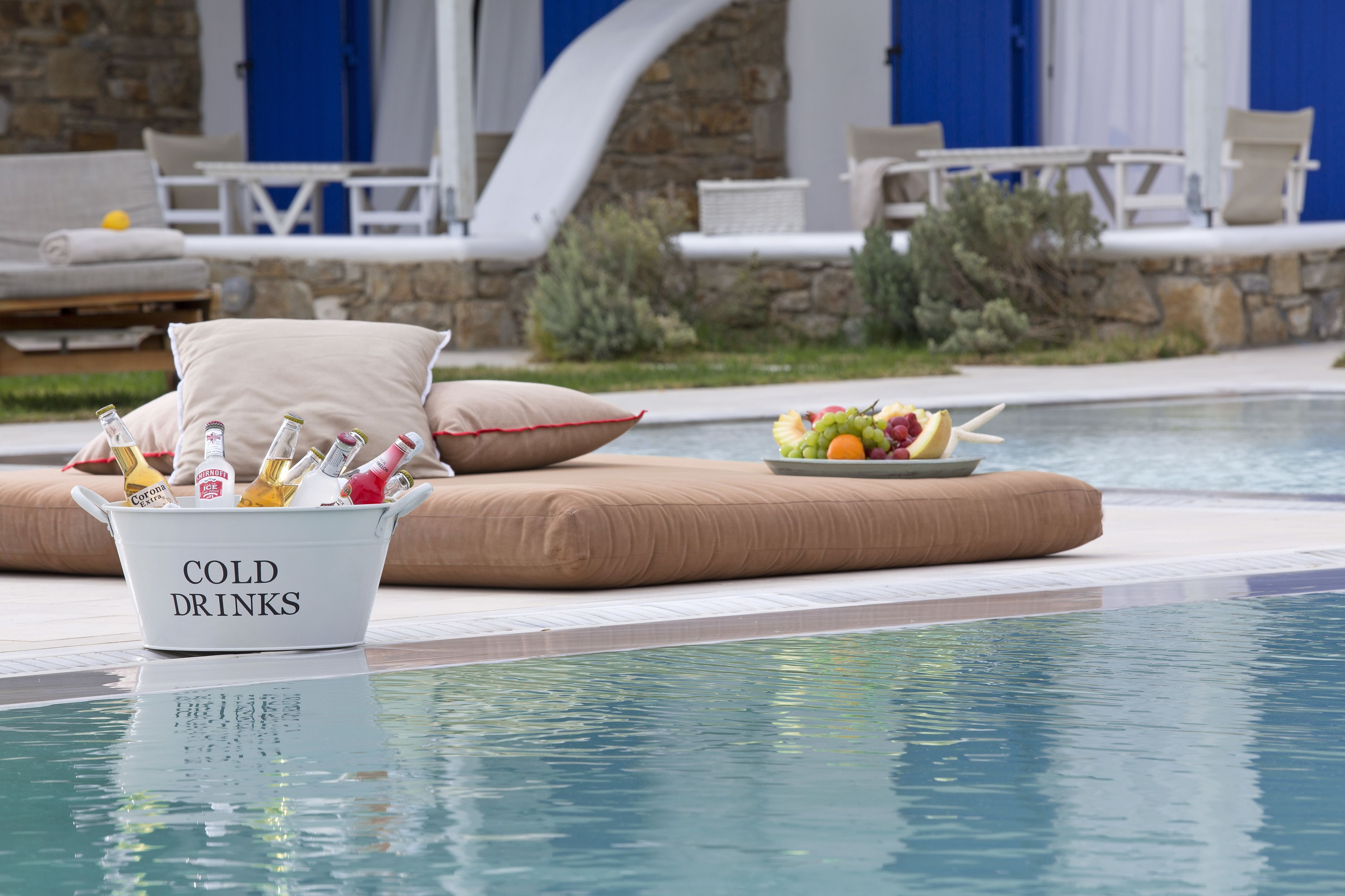 A poolside area with a bucket labeled "COLD DRINKS" filled with bottles, a fruit bowl, and a lounge chair with cushions.