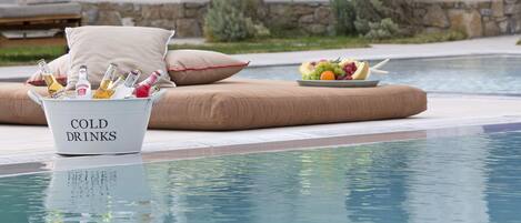 A poolside area with a bucket labeled "COLD DRINKS" filled with bottles, a fruit bowl, and a lounge chair with cushions.