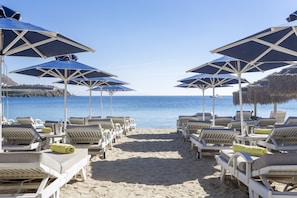 A beachside lounge area with rows of sunbeds and umbrellas, a clear view of the sea, and a thatched-roof structure in the background. - Villa Del Sol (Mykonos)