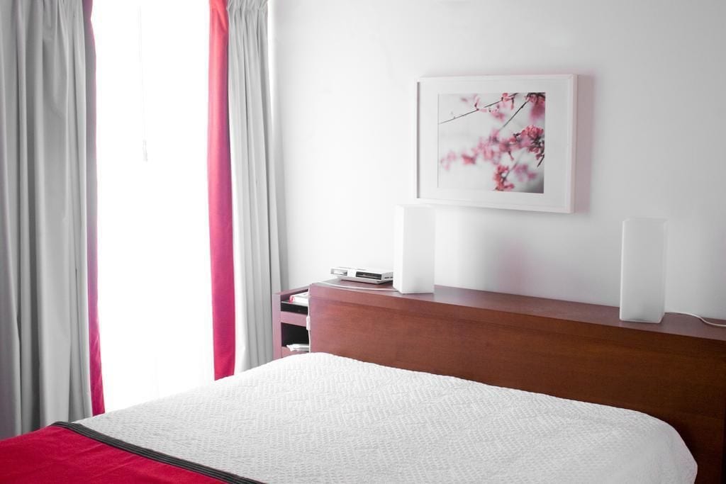A hotel room with a bed, a wooden nightstand, a framed picture on the wall, and a window with curtains.