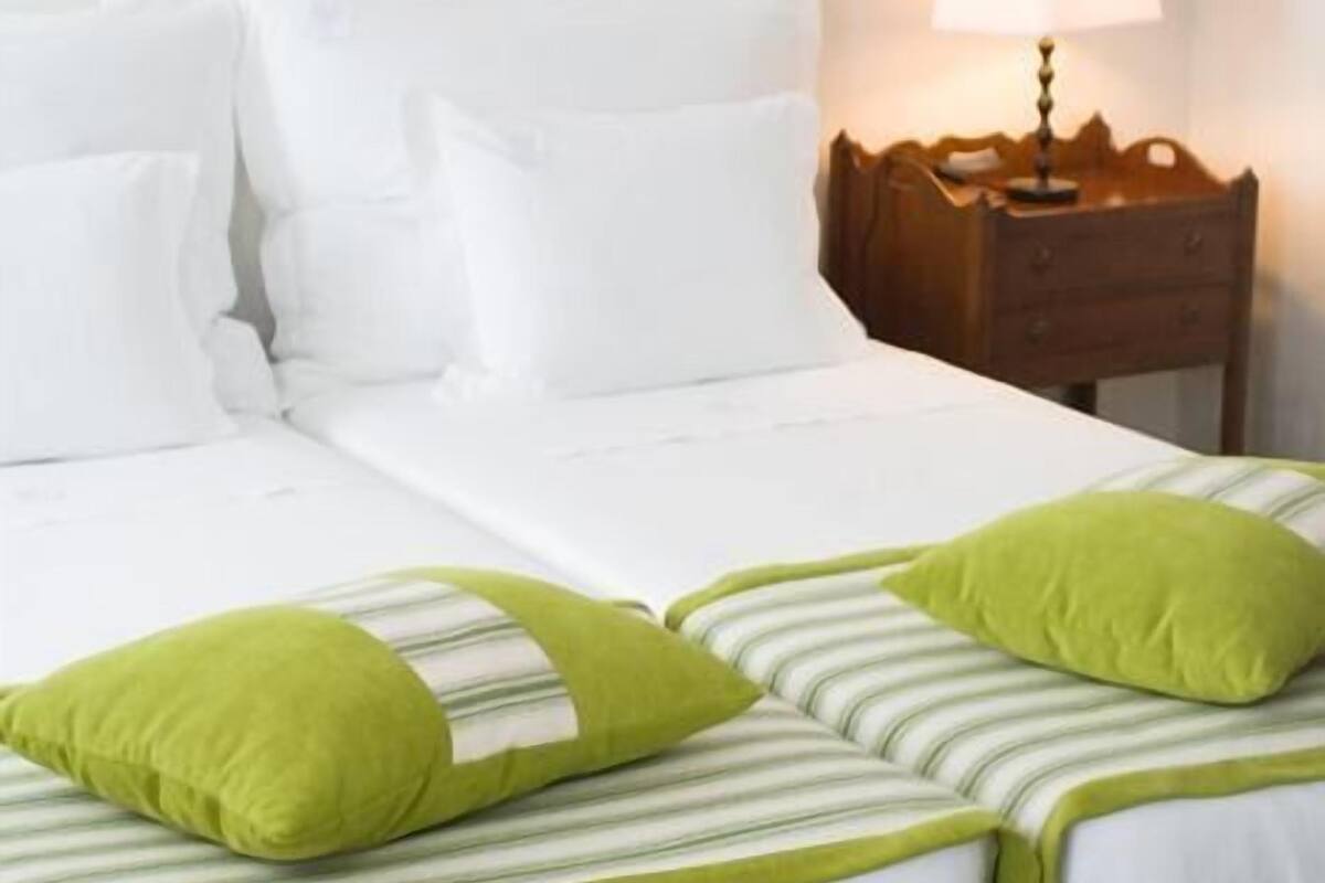 A neatly made bed with white bedding and green pillows, a wooden nightstand with a lamp, and a plain white wall.