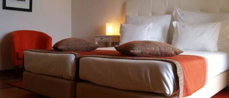 A hotel room with a double bed, two pillows, a nightstand with a lamp, a red chair, and a framed picture on the wall.