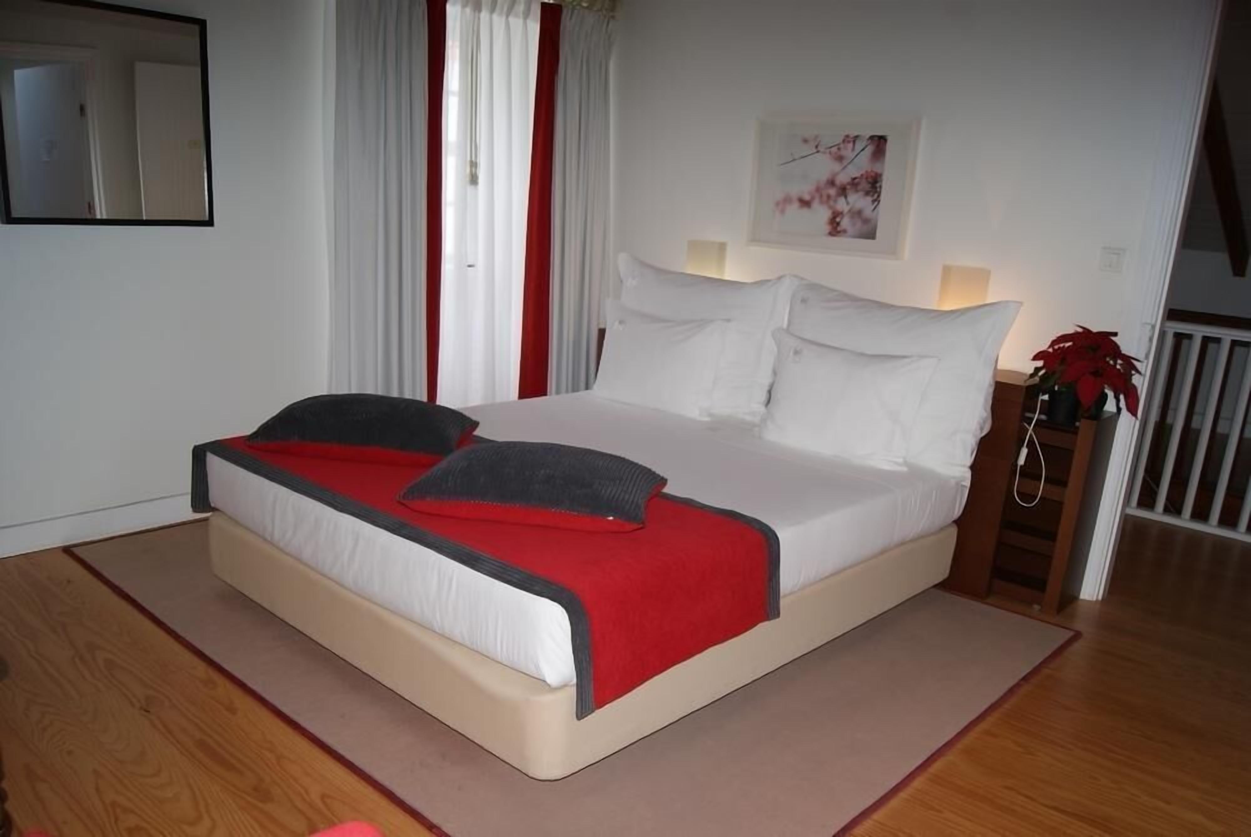Un lit bien fait, avec du linge de lit blanc et des touches de rouge, une table de chevet avec une lampe, et une fenêtre ornée de rideaux blancs.