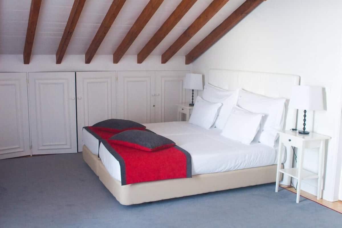 A bedroom with a bed, white walls, wooden beams, and a nightstand with a lamp.