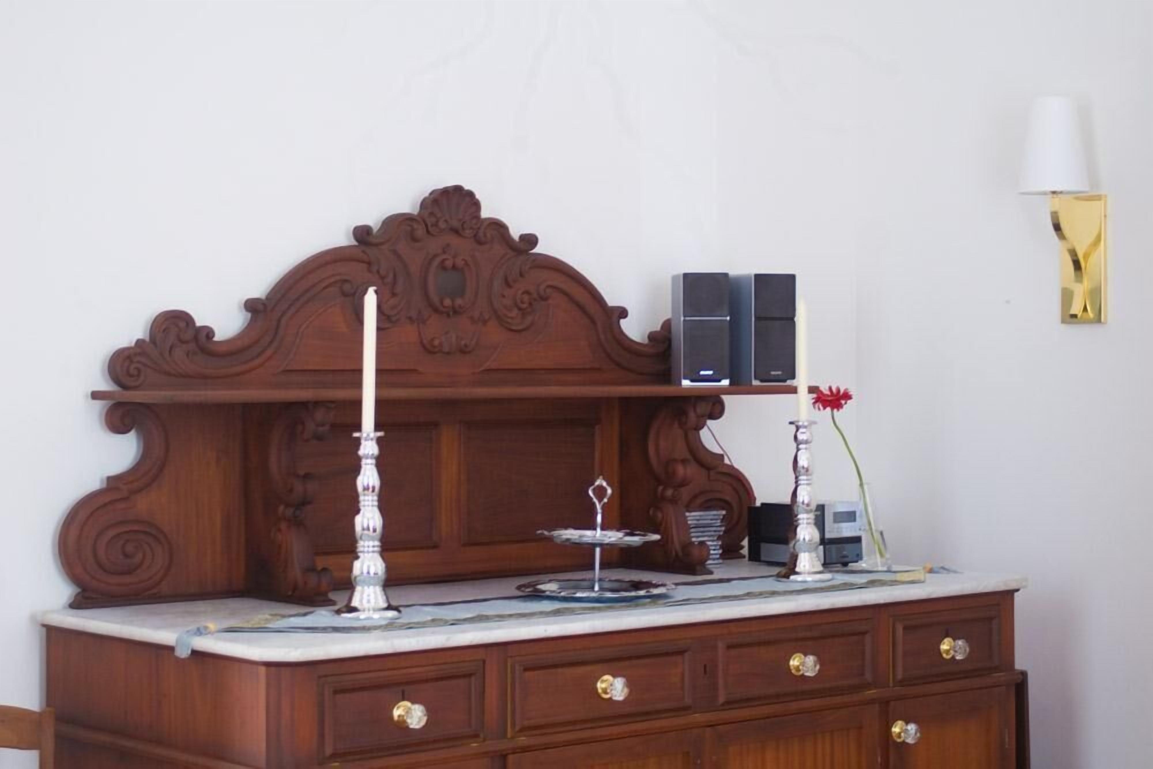 Uma cômoda de madeira com tampos de mármore, duas velas, uma pequena mesa com castiçal e um abajur fixado na parede.